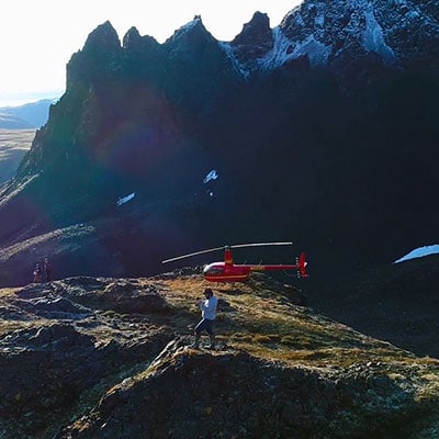 Aerial view of helicopter sitting on mountain ridge with pilot standing beside it.